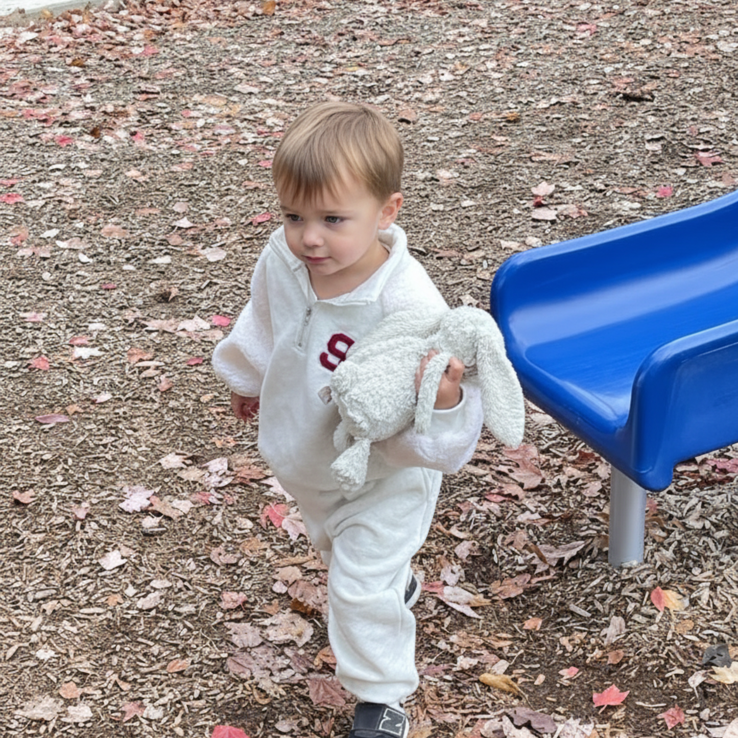 Child in a heather gray outfit holding a stuffed animal on a playground with a blue slide.